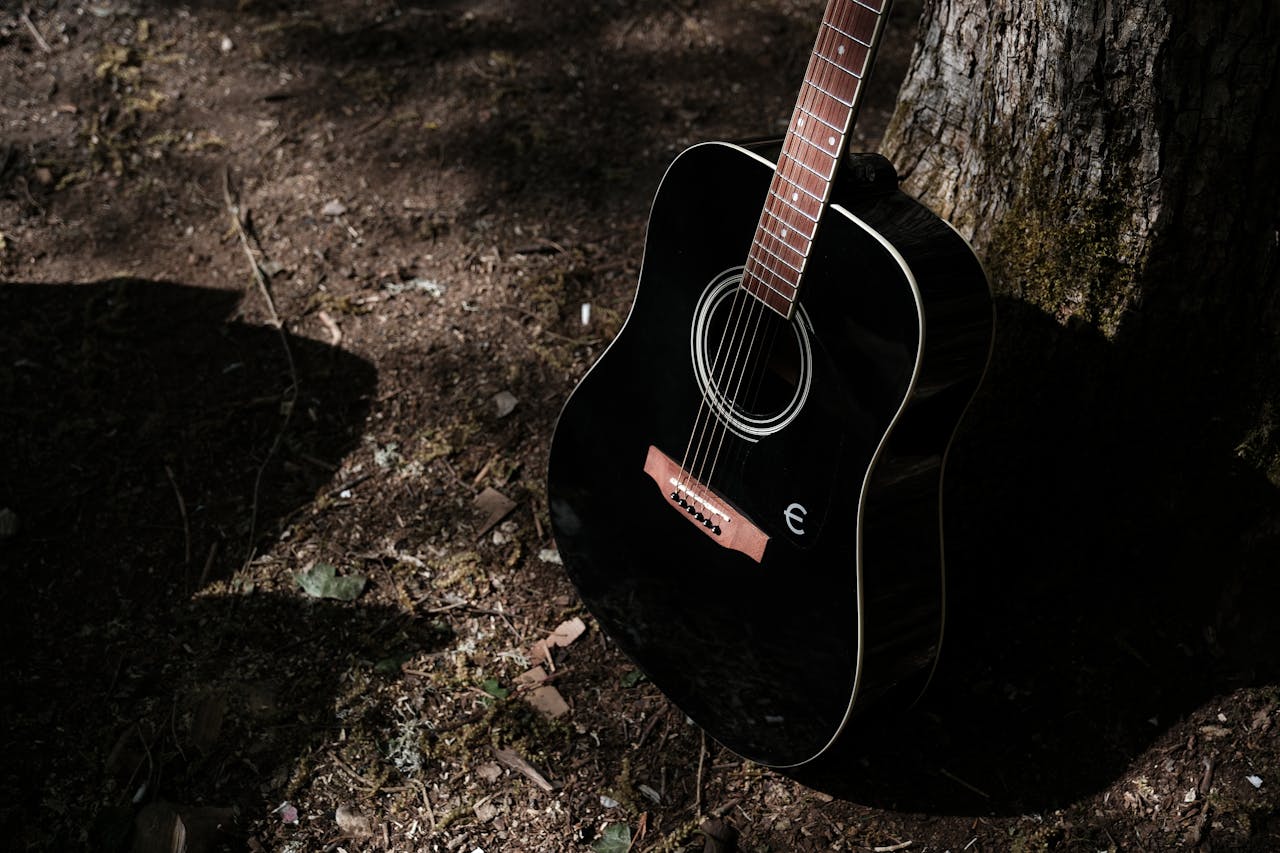 A black acoustic guitar rests against a tree in a forest, showcasing its elegant design.